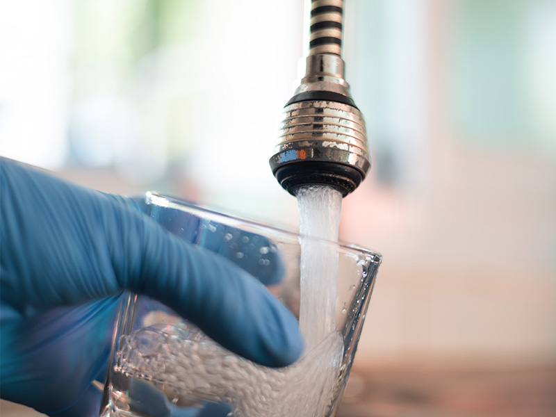 Gloved hand holding glass collecting water sample from faucet.
